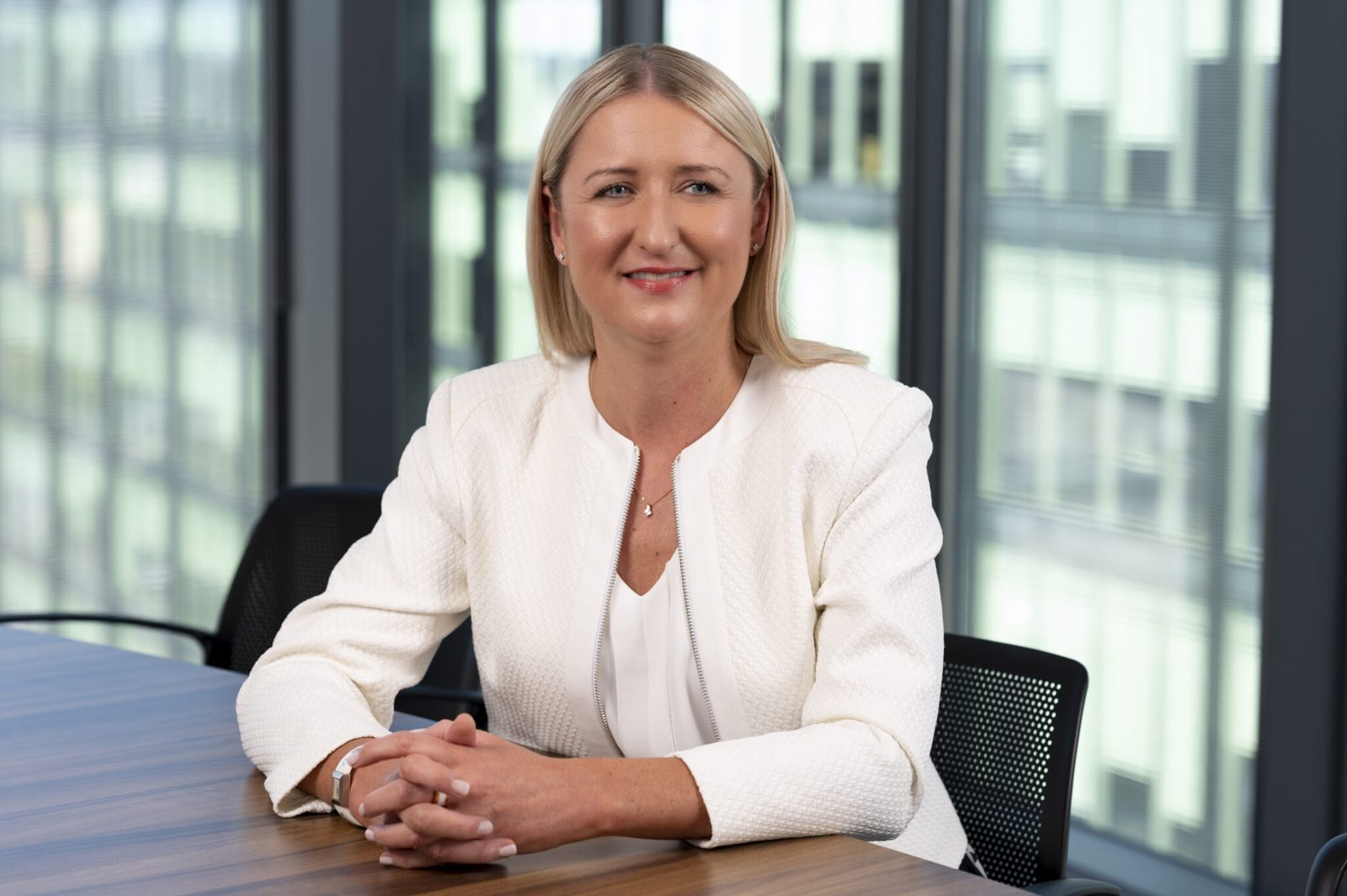 Woman in white blazer seated at table, smiling, office window background