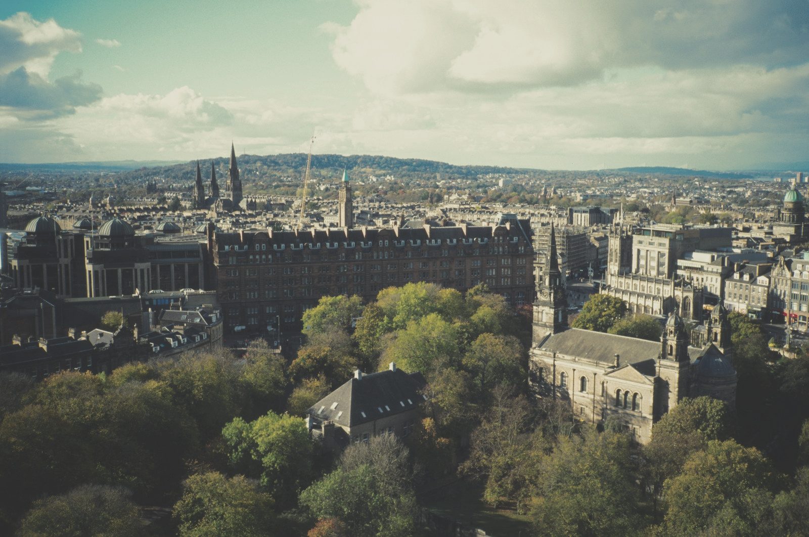 Elevated daytime view of Edinburgh skyline with historic stone buildings, spires, trees, and hills