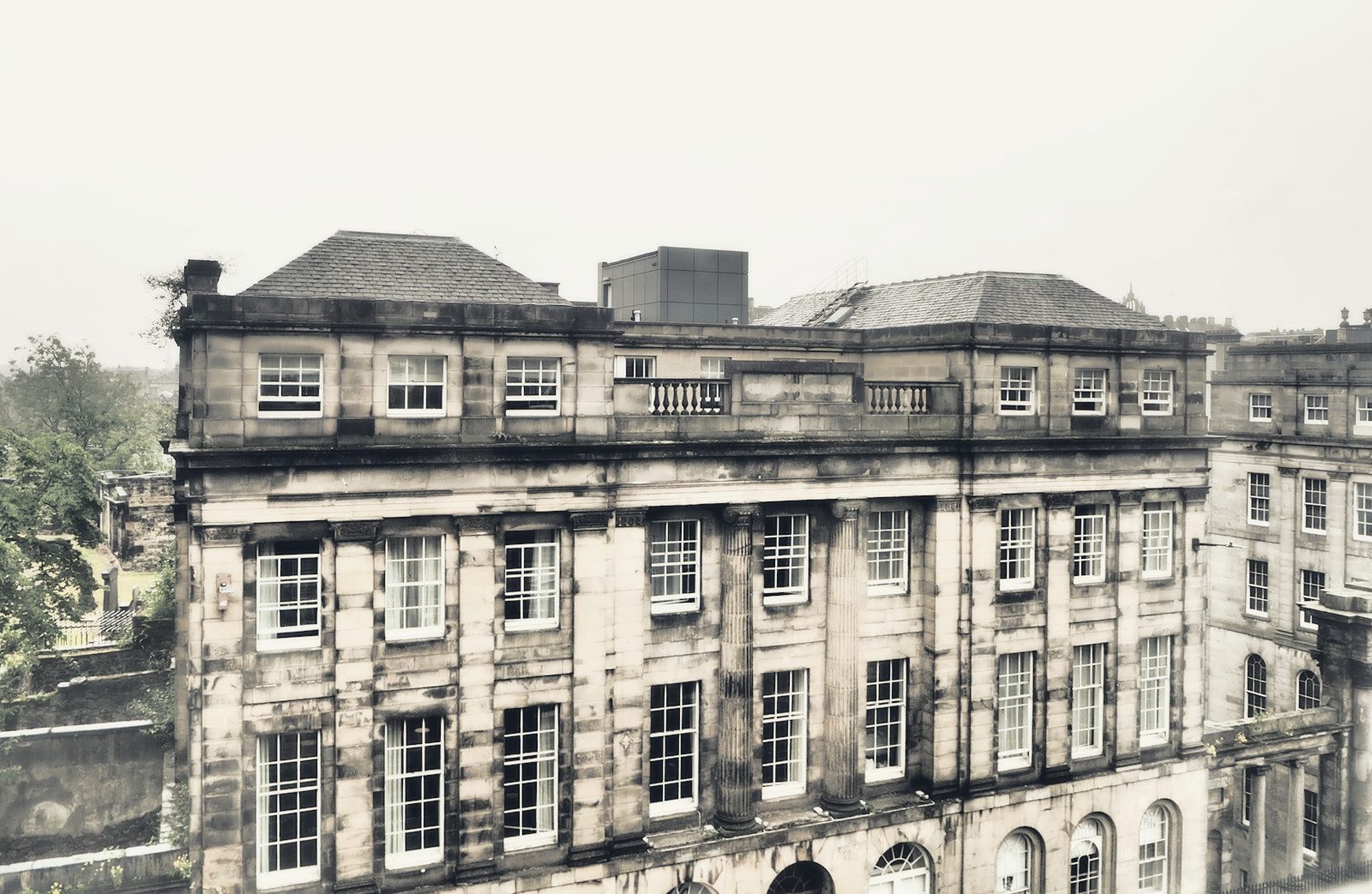 Weathered stone neoclassical building facade with columns, sash windows, and sloped roof under grey sky